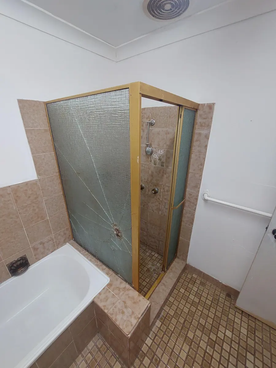 Damaged gold-framed shower screen with cracked glass in a tiled bathroom